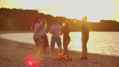 The-students-are-dancing-around-bonfire-on-the-beach-with-beer.-They-are-talking-to-each-other-at-sunset-and-enjoying-the-summer-evening-on-the-river-coast.